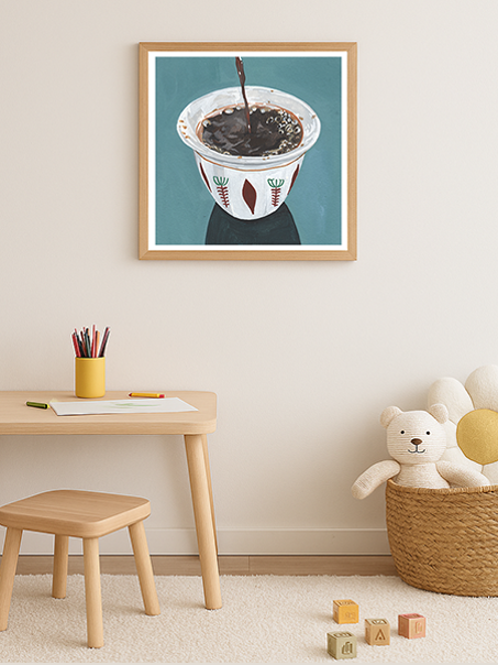 A framed wall art print showing a white bowl with a dark liquid and colorful illustrations, displayed in a styled children's room with a wooden desk, stool, basket, and stuffed toys.