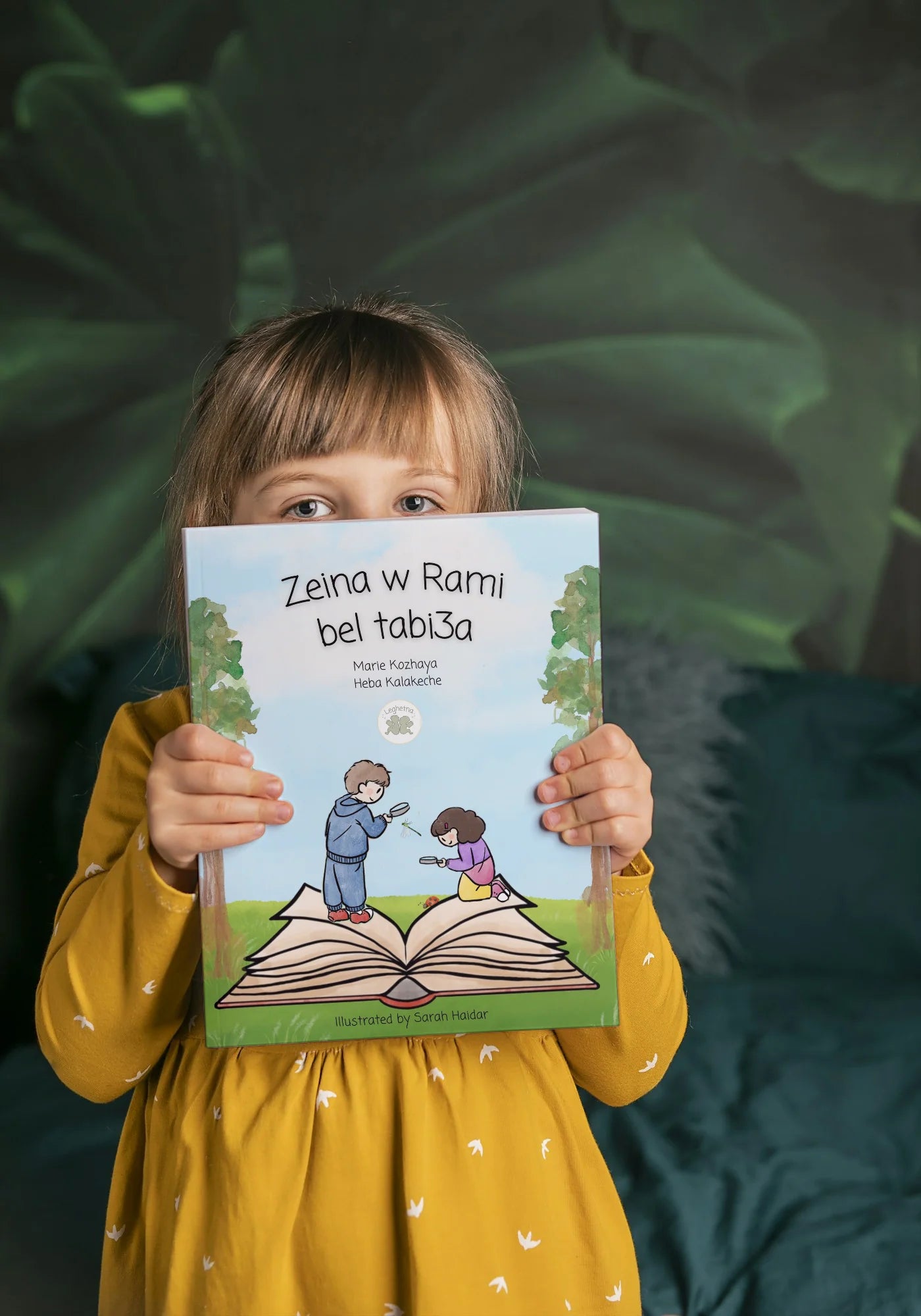 Child holding a book titled 'Zeina w Ramji bei tabisa' with a green leafy background