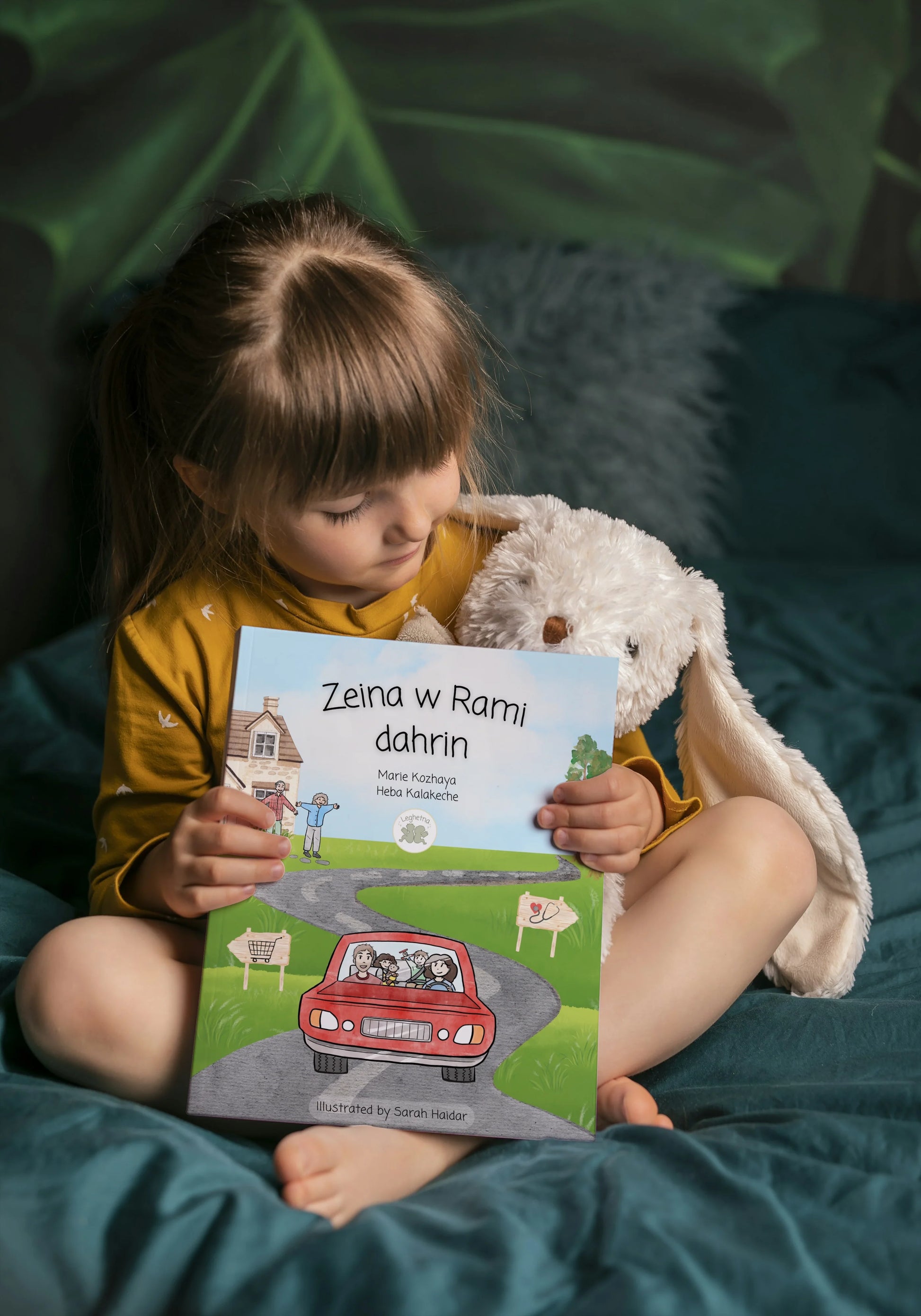 Child holding a book titled 'Zeina w Rami dahrin' with a soft toy on a green cushion.