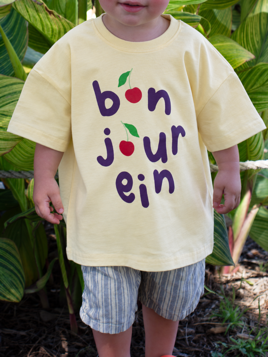 A toddler wearing a pale yellow t-shirt with a 'bonjour' graphic made of cherries, standing outdoors in front of green tropical leaves.