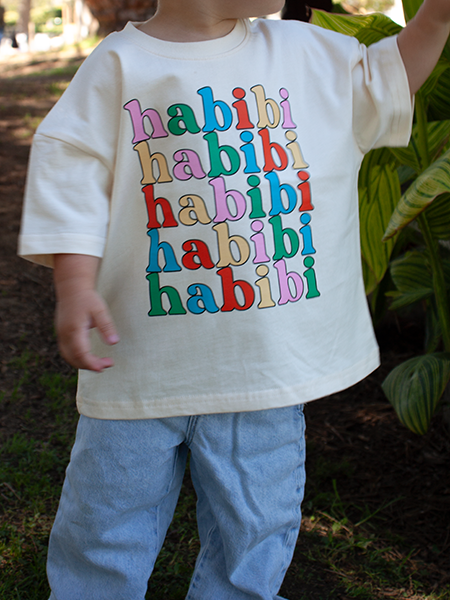 A child wearing a cream-colored t-shirt with the word 'habibi' repeated in colorful bubble letters, paired with light blue jeans, standing outdoors in front of green plants.