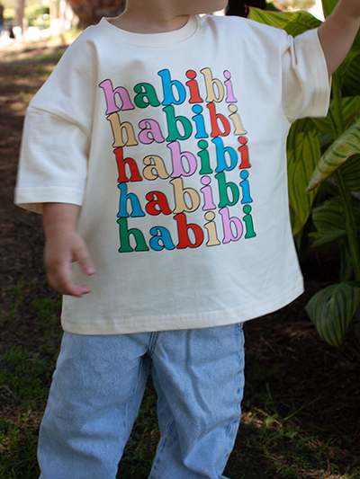 A child wearing a cream-colored t-shirt with the word 'habibi' repeated in colorful bubble letters, paired with light blue jeans, standing outdoors in front of green plants.