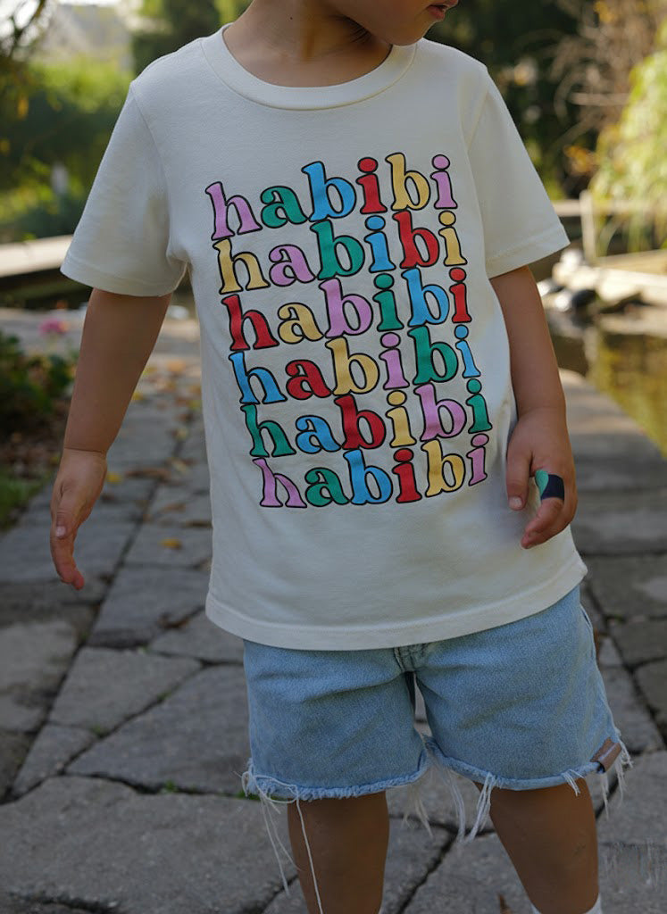 Child wearing a white t-shirt with 'habibi' printed in colorful letters outdoors.