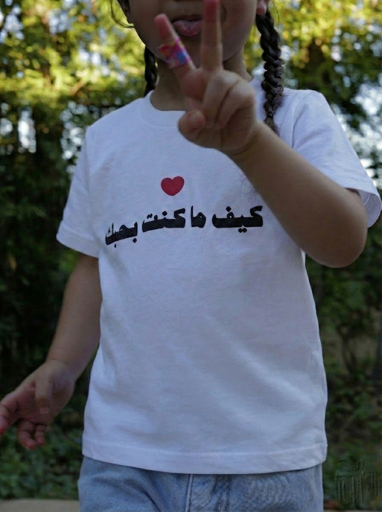 Child wearing a white t-shirt with Arabic text and a heart symbol, making a peace sign outdoors.