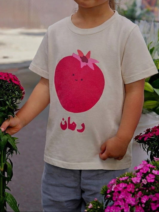Child wearing a t-shirt with a red pomegranate design and Arabic text, standing among flowers.