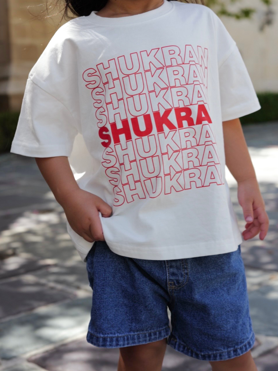 A child wearing a white oversized T-shirt with the word 'SHUKRAN' repeated in red and light pink outline text, paired with blue denim shorts, photographed outdoors on a paved walkway.