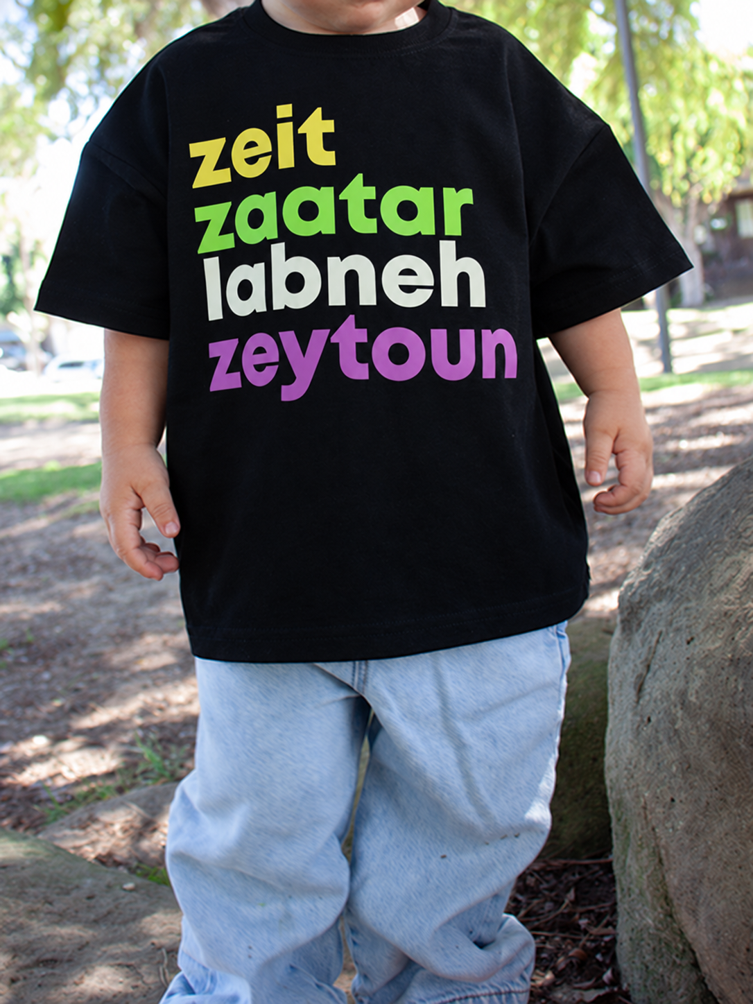 A child wearing a black graphic t-shirt with the words 'zeit zaatar labneh zeytoun' printed in yellow, green, white, and purple, paired with light blue jeans, standing outdoors next to a large rock.